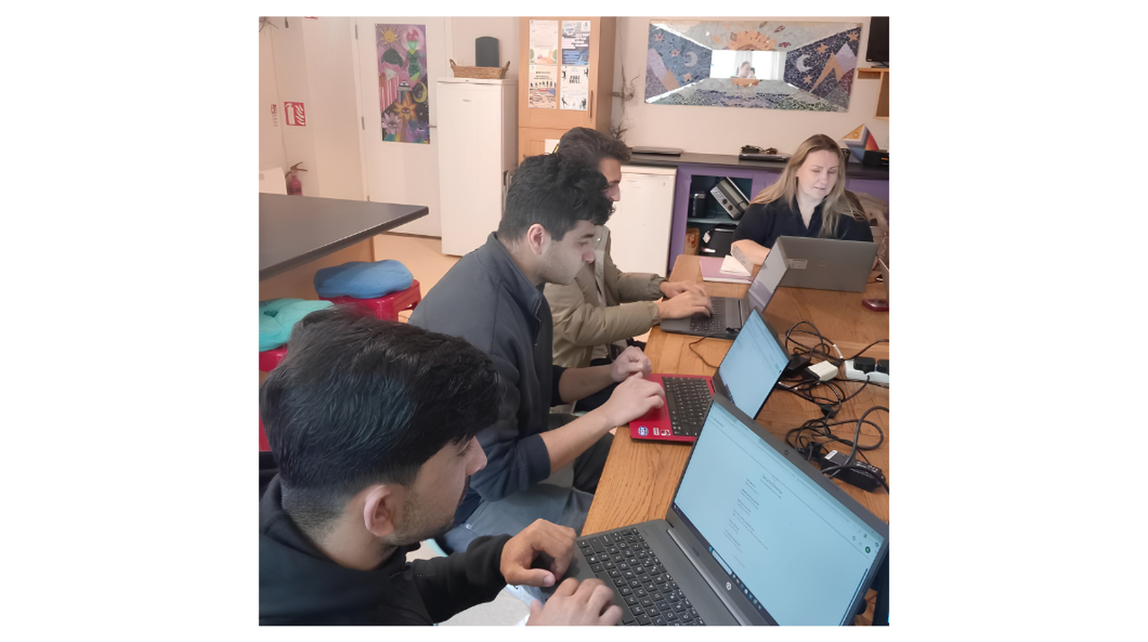 Four people working on laptops at a wooden table in a room with colorful posters and a mirror on the wall.